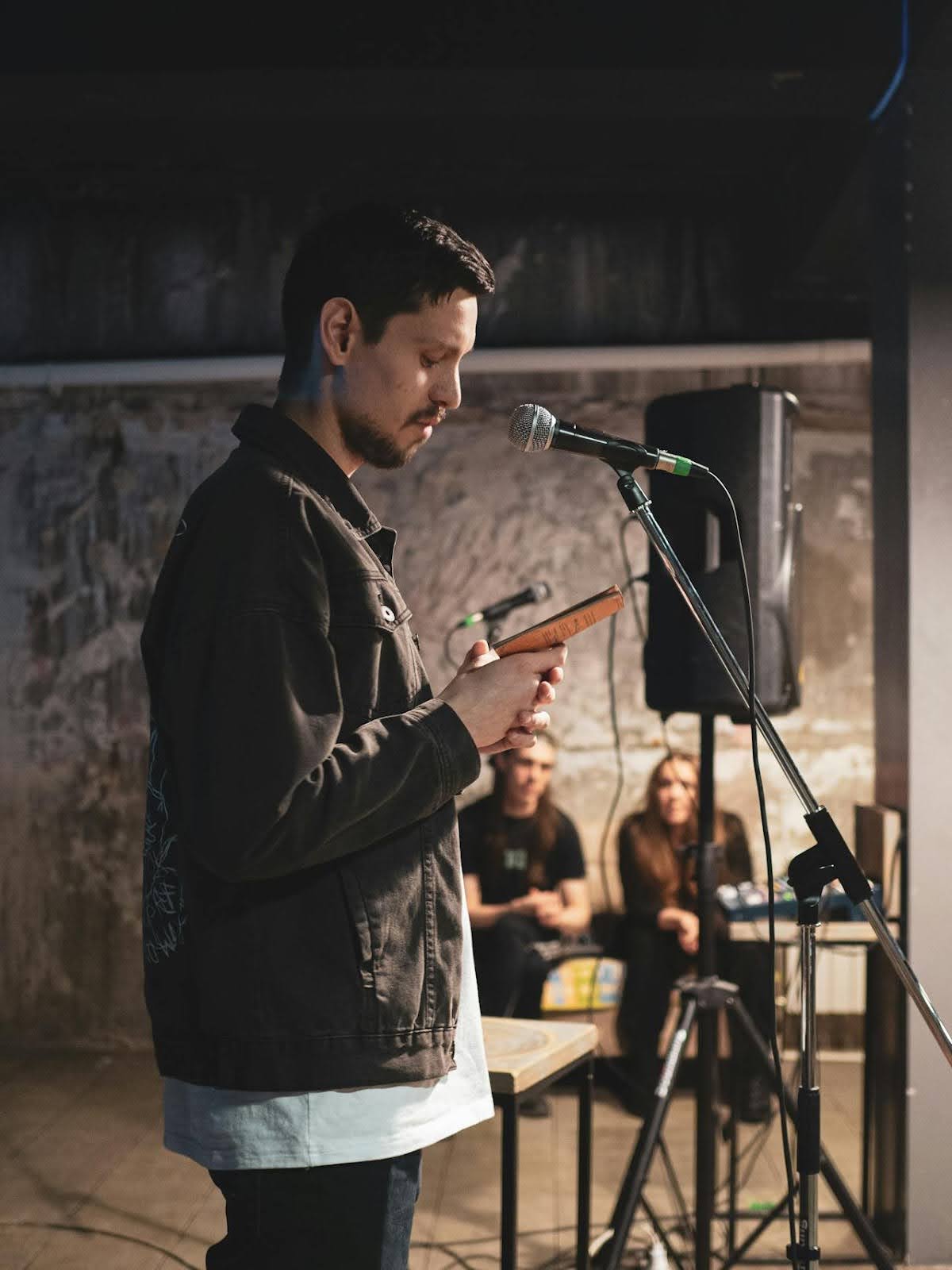 A person in a denim jacket reads from a book at a microphone on stage. Two people sit in the background, listening.